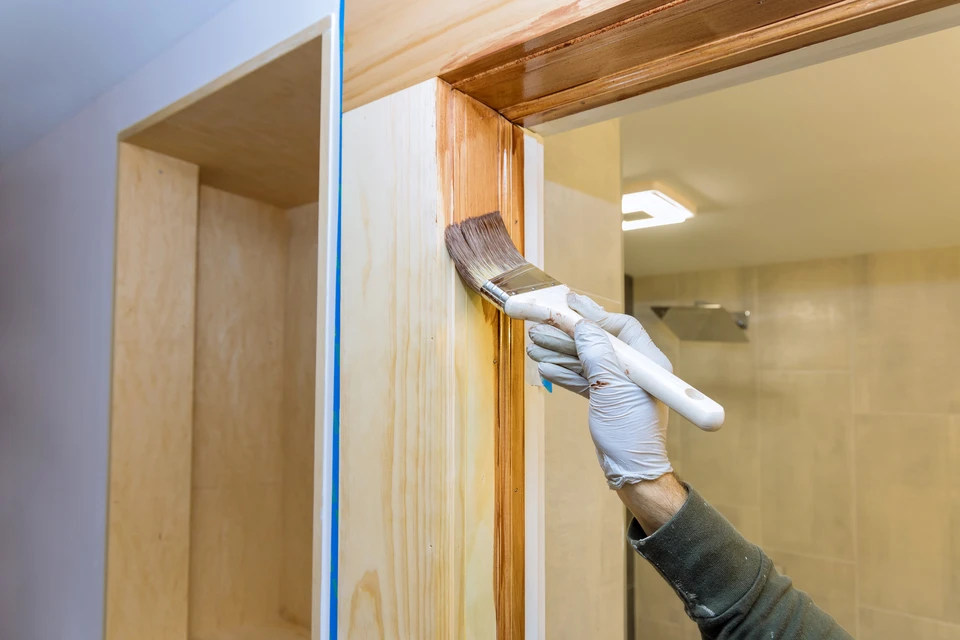 Intérieur résidentiel, main gantée appliquant vernis ou lasure sur boiserie de porte avec pinceau plat